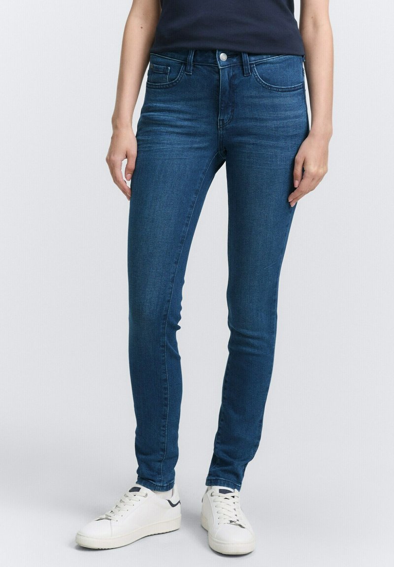 Jeans ajustados de color azul, hechos de denim con una textura suave, que presentan un cierre frontal con botón y bolsillos laterales, combinados con zapatillas blancas.