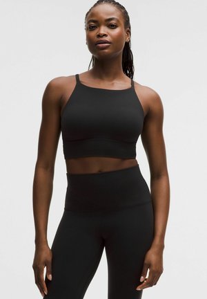 Atletisk kvinde står iført en sort croppet tanktop og højtaljede sorte leggings mod en ensartet lys baggrund.