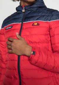 Rød og marineblå pufjakke med lynlås foran, vandrette baffler, broderet logo og kontrasterende hvide detaljer langs skuldrene.