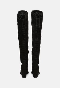 Pár černých semišových kozaček po kolena s blokovými podpatky, detaily šněrování na zadní straně a postranními zipy, zobrazené zezadu na bílem pozadí.