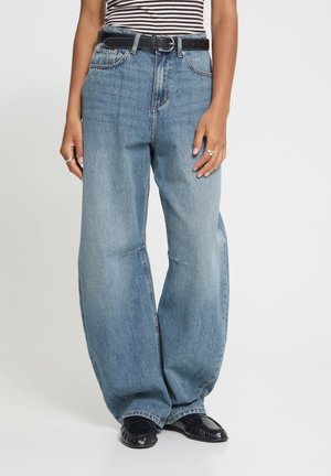 Personne portant un jean bleu à jambes larges, une ceinture noire, des chaussures noires, un haut sans manches rayé et des bagues, debout devant un fond uni.