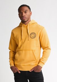 Felpa gialla realizzata in un tessuto morbido con tasca frontale e patch con logo. Il design include cappuccio con coulisse e polsini a coste.