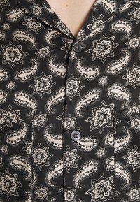 Chemise noire à manches courtes ornée d'un motif cachemire et floral beige, avec des boutons au col et sur la poche, fabriquée dans un tissu lisse.