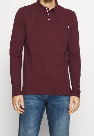 Mand iført en langærmet bordeaux polo skjorte med frontlomme og blå jeans, stående mod en ensfarvet baggrund.