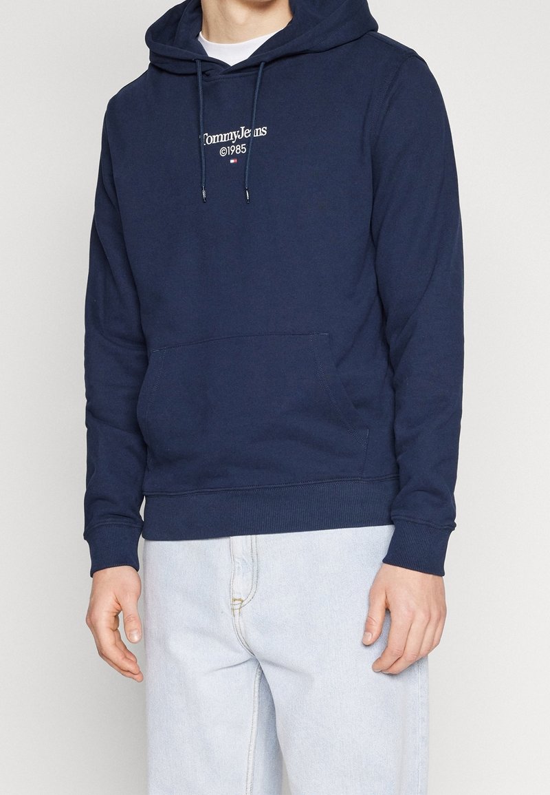 Námořnická modrá mikina s kapucí, která má klokankovou kapsu a šňůrku na stažení. Na hrudi je vyšitý bílý logo "Tommy Jeans". Měkký bavlněný materiál.