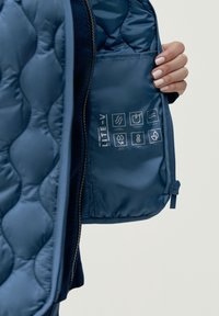 Interior de chaqueta acolchada azul con un bolsillo con cremallera que contiene instrucciones de cuidado, íconos para el mantenimiento y el texto "LITE-V" impreso en la tela.