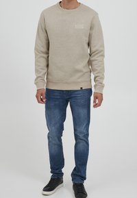 Béžová bavlněná mikina s kulatým výstřihem a žebrovanými manžetami, na hrudi malý bílý logo. Ve spojení s modrými džínami a černými teniskami.