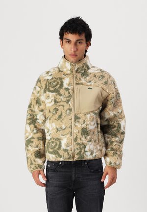 Fleece jacket with a floral pattern in beige and green, featuring a zip-up front and a tan chest pocket with branding.