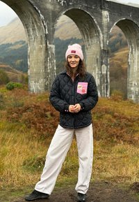 Mladá žena s růžovou čepicí a černou prošívanou bundou stojí na travnaté cestě poblíž kamenného mostu s obloukem v kopcovité krajině.