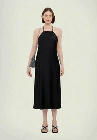 Sieviete stāv melnā bezpiedurkņu halter midi kleitā, turot mazu melnbaltu šaha rūtiņu somu, ģērbusies melnās siksniņu kurpēs.