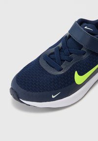 Chaussure de sport bleu marine avec tige en mesh, accents gris tonal et un logo Nike jaune vif. Dotée d'une fermeture Velcro et de lacets. Semelle blanche.
