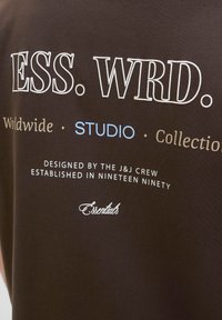 Brown cotton T-shirt featuring white and light blue text, including "ESS. WRD." and "STUDIO," with a smooth texture and relaxed fit.