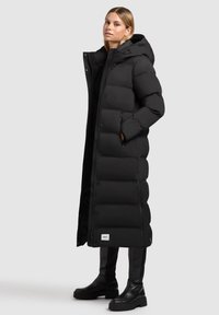 Abrigo largo negro tipo plumón con capucha, con secciones acolchadas, cremallera frontal y bolsillos laterales. Llevar encima de pantalones ajustados negros y botas.