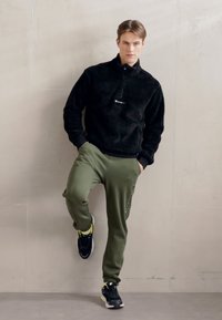 Sort fleece sweatshirt med halv-lynlås, olivengrønne tapered sweatpants og sorte sneakers med gule detaljer, stående mod en neutral væg.