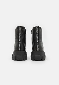 Bottes noires épaisses avec semelles crantées épaisses, fermeture éclair arrière et languettes verticales affichant le texte "Steve Madden", vues de dos.