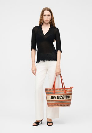 Sieviete ar viļņainiem matiem, tērpusies caurspīdīgā melnā topā, baltās biksēs un melnās atvērtām purngalu kurpēm, turot svītrainu Love Moschino somu ar oranžām rokturiem.