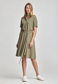 Abito camicia verde oliva con maniche corte, apertura con bottoni, cintura in vita e texture morbida. Abbinato a sneaker bianche per un contrasto.