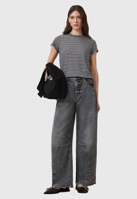 T-shirt rayé noir et blanc, pantalon en denim gris lâche avec des ourlets effilochés, et chaussures noires sans lacets. Tenant un sac noir avec du texte.