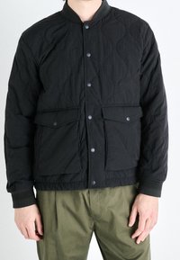 Zwarte gewatteerde jas met een knoopsluiting, ribgebreide manchetten en twee borstzakken. Gestructureerde stof, lichtgewicht ontwerp en een ontspannen pasvorm.