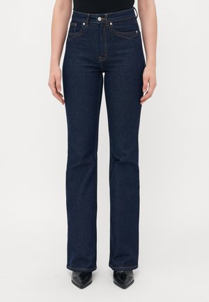 Woman wearing dark blue high-waisted straight-leg jeans and black pointed-toe shoes, standing against a plain light background.