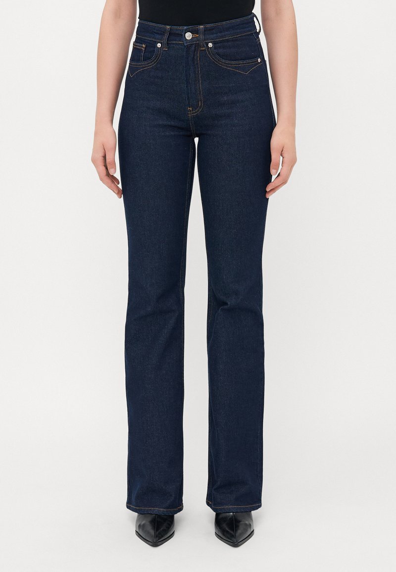 Woman wearing dark blue high-waisted straight-leg jeans and black pointed-toe shoes, standing against a plain light background.