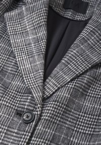 Villa villasekoitemateriaalia, jossa on musta-valkoinen hounds-tooth-kuvio, läppälaput ja yksittäinen musta nappikiinnitys. Vuorattu sisus.