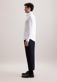 Man in profiel staande in een wit overhemd met lange mouwen, zwarte pantalon, zwarte sokken en zwarte nette schoenen, voor een effen achtergrond.