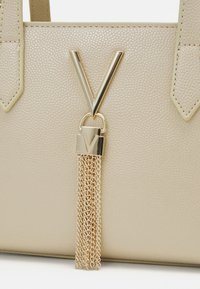 Beigemelerad handväska med ett guld V-format accent och en kedjehängsel med en ingraverad låsdetalj.