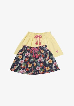Tricou alb cu mânecă scurtă, având un design cu flamingo roz și grafică cu bretele, asociat cu pantaloni scurți galbeni deschiși și adidași albi.