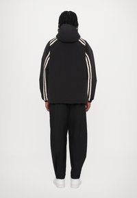 Črna jopa s kapuco z bež črtami ob ramenih, kombinirana z ohlapnimi črnimi hlačami in belimi supergami, pogled od zadaj.