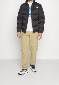 Czarna kurtka puchowa z zamkiem z przodu, w połączeniu z beżowymi spodniami cargo z zamkami na bokach. Czarno-białe sneakersy dopełniają stylizację.
