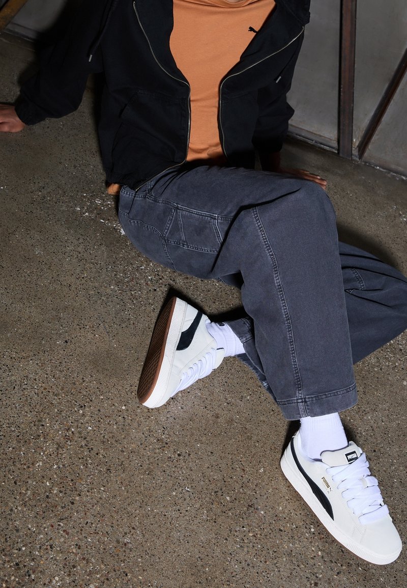 Personne portant des baskets blanches Puma avec une bande noire, des chaussettes violet clair, un pantalon gris foncé, une veste noire et une chemise orange, assise sur un sol en béton.