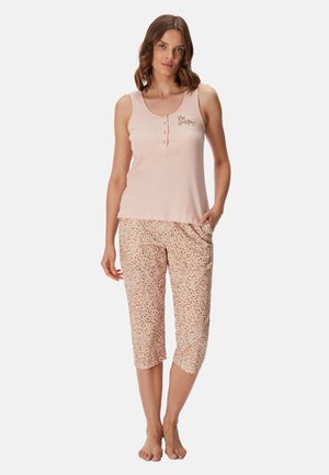 Vrouw die een lichtroze mouwloze top met knopen en tekst "Be Happy" draagt, gecombineerd met cropped pyjamabroek met luipaardprint, staat blootsvoets.