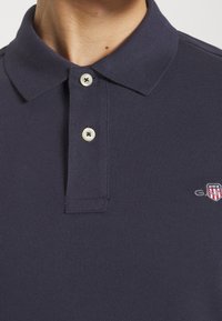 Marinblå polo t-shirt i en texturerad väv, med två knappar och en liten logotyp med röda, vita och blå detaljer på vänster bröst.