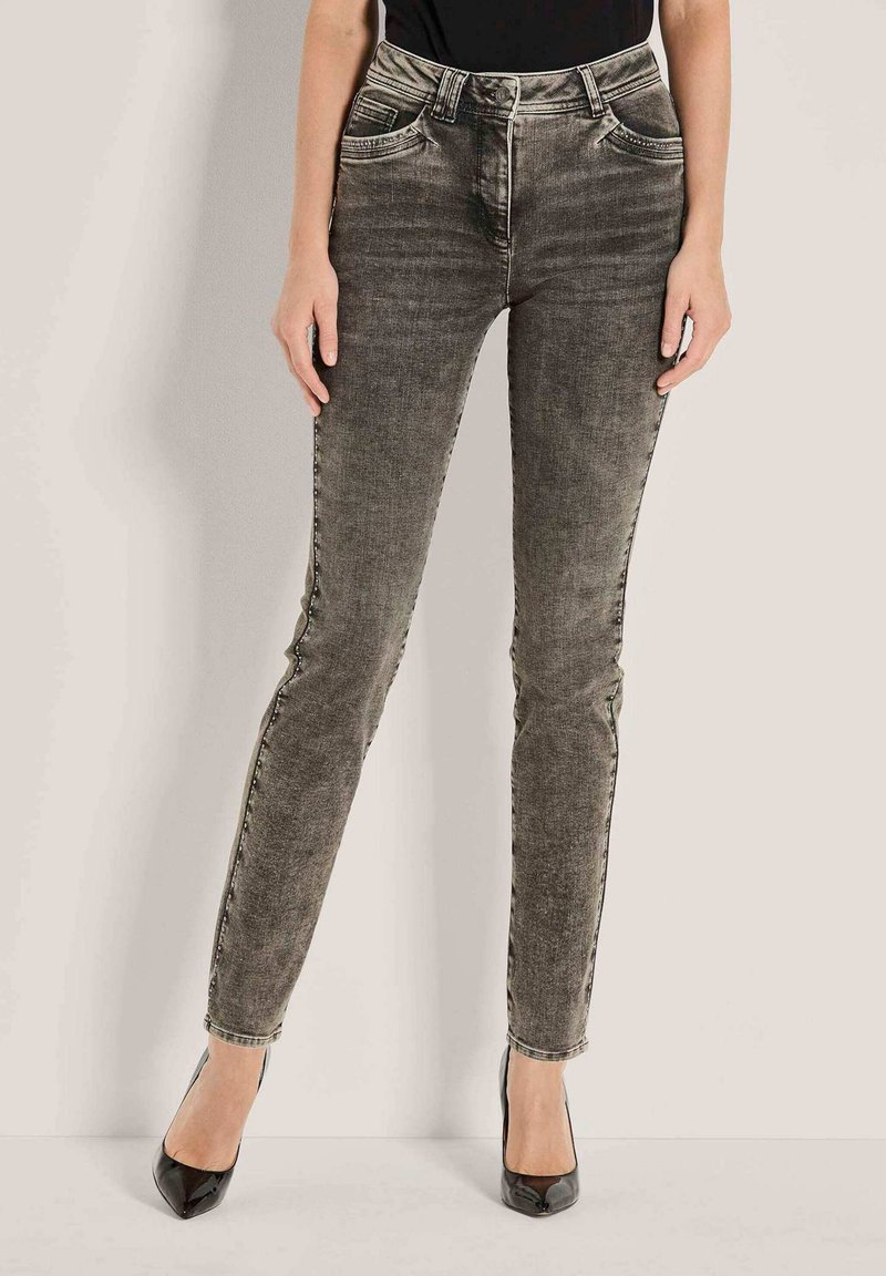 Vrouw in slim-fit vervaagde zwarte jeans met gestippelde zijnaad en zwarte puntige hakken, staande tegen een lichte muur.