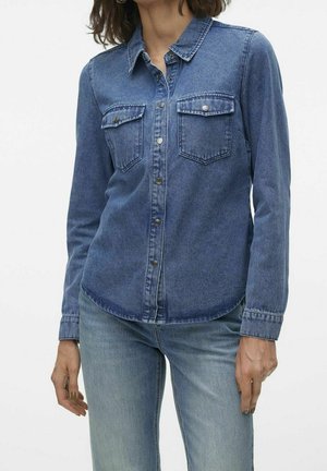 Femme portant une chemise en jean bleue à boutons avec des poches poitrine et un jean bleu clair sur fond blanc uni.