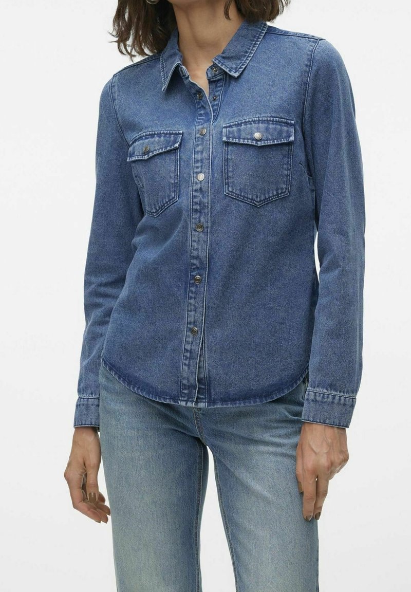 Femme portant une chemise en jean bleue à boutons avec des poches poitrine et un jean bleu clair sur fond blanc uni.