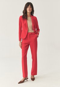 Žena stojící v červeném dámském obleku s béžovým topem a hnědými slingback podpatky na pozadí světlé barvy.