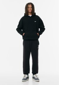 Zwarte hoodie met een voorzak en logo, gecombineerd met zwarte sweatpants. Sneakers hebben een grijs en witte patroon. Katoenen materiaal, relaxte pasvorm.