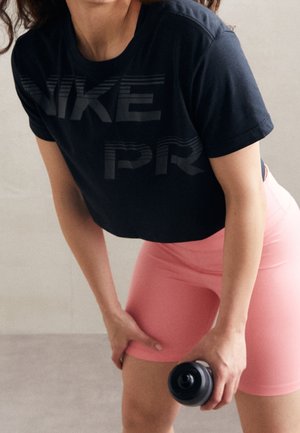 Osoba w czarnej koszulce Nike Pro i różowych szortach trzyma czarną butelkę na wodę, pochylając się do przodu z jedną ręką opartą na udzie.