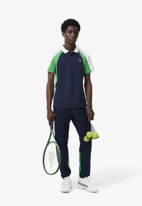 Polo navy con dettagli verdi e bianchi, presenta un colletto e due bottoni, abbinato a pantaloni navy e sneakers bianche, mentre tiene una racchetta da tennis e delle palline.