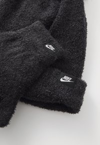 Crni puhasti pleteni šešir i rukavice s crnim Nike oznakama. Teksturirana tkanina izgleda toplo i mekano, s jednostavnim dizajnom bez uzoraka.