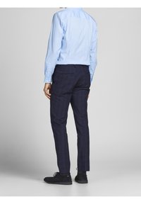 Ljusblå långärmad skjorta i kombination med mörkblå rutiga byxor och svarta mockaskor, sedd bakifrån. Knappar och fickdetaljer synliga.