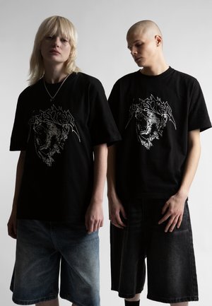 Dvaja stojaci ľudia oblečení v čiernych oversized tričkách s grafikou revúceho medveďa a tmavých krátkych nohaviciach do kolien na jednoduchom pozadí.