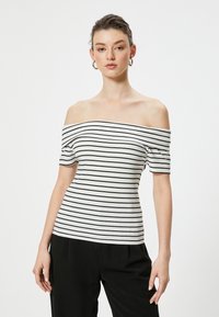 Off-the-shoulder top med ribbet tekstur, prydet med sorte vandrette striber på en hvid baggrund. Korte ærmer, tætsiddende design.
