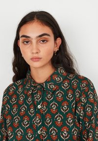 Jeune femme avec de longs cheveux brun foncé portant un blouse verte avec des motifs floraux orange et de petites boucles d'oreilles en or contre un fond blanc.