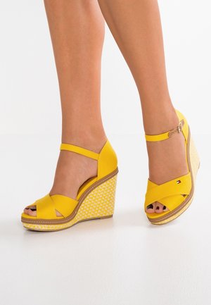 Gule wedgesandaler med et crisscross-stroppedesign, åben tå og et mønstret hvidt mellemsål. Justerbar ankelrem inkluderet.