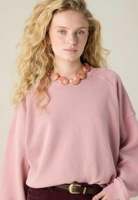 Femme blonde avec des cheveux bouclés, portant un gros pull rose, un grand collier de perles roses et un pantalon sombre avec une ceinture marron.