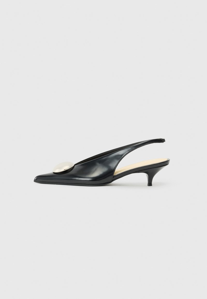 Zapato de charol negro tipo slingback con punta afilada, tacón bajo y un gran acento metálico en la parte frontal. Textura suave, diseño minimalista.