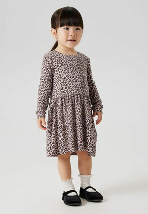 Långärmad klänning i ljus rosa med svart leopardmönster, sammanknuten midja och utsvängd kjol; kombinerad med vita volangstrumpor och svarta skor.